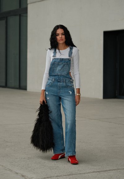 Unos overoles de mezclilla con detalles desgastados, combinados con una blusa blanca de mangas largas, zapatos rojos y un bolso de piel de pelo negra texturizada.