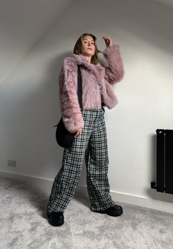 Jachetă din blană sintetică de culoare roz deschis, pantaloni largi cu carouri negre și pantofi masivi negri, acompaniată de o geantă de umăr neagră. Textura și modelul sunt vizibile.