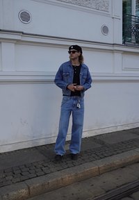 Denim jacket and wide-leg jeans in light blue. Black henley shirt underneath, paired with a black cap and sunglasses. Keyring detail visible.