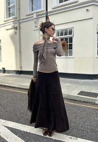 Brūns tops bez uzlādēm, pelēks šalle, gara brūna zamša svārki un spīdīgas burgundijas krāsas kurpes. Stāv uz ielas ar ēkām fonā.