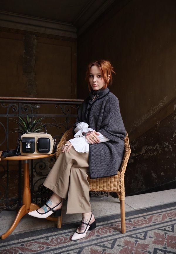 Tânără cu păr roșcat stă pe un scaun de ratan, purtând o pelerină gri, pantaloni bej și pantofi albi, lângă o masă cu o geantă bej și neagră.