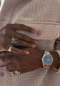 Des mains à la peau foncée avec des bagues en argent et une montre-bracelet reposent sur une veste en laine lavande avec un bouton et un texte brodé "Fournie."