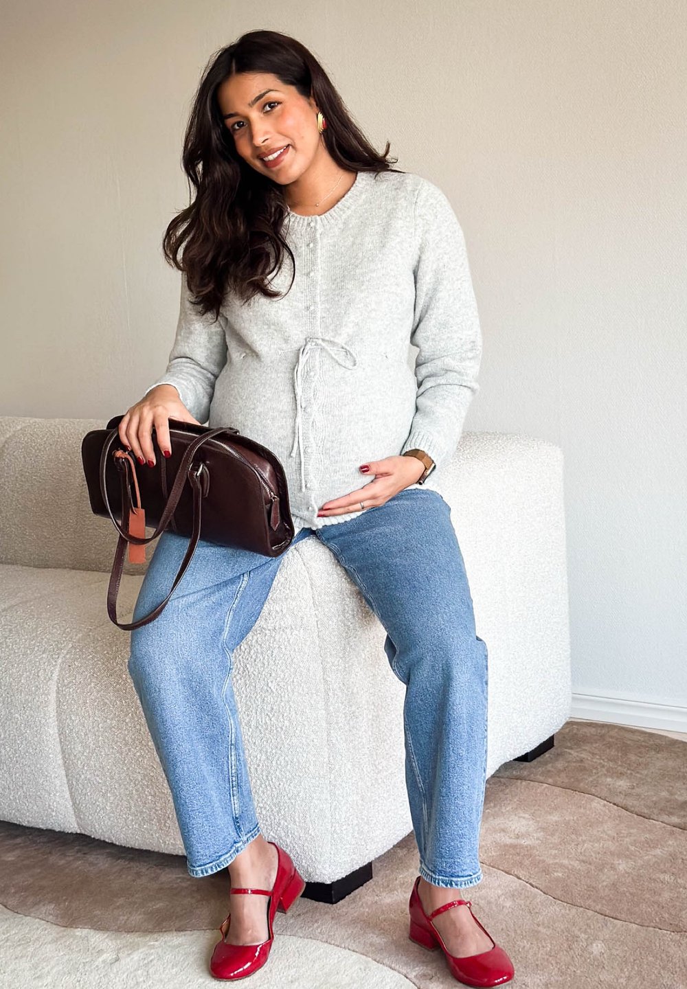 Femme enceinte en pull gris et jean assise sur un canapé beige, tenant un sac à main marron foncé, portant des chaussures rouges, souriant à la caméra.