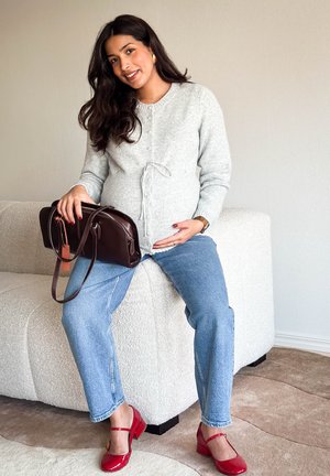 Mujer embarazada con suéter gris y jeans sentada en un sofá beige, sosteniendo un bolso marrón oscuro, con zapatos rojos, sonriendo a la cámara.