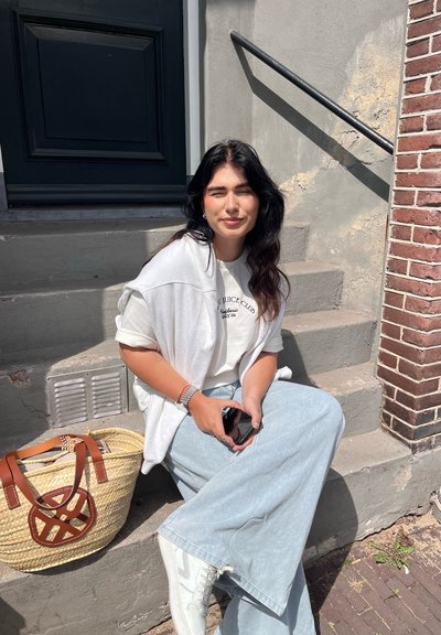 Suéter blanco sobre camiseta gris clara, jeans anchos de color azul claro, zapatillas blancas y un bolso de mimbre con correas y detalles marrones.