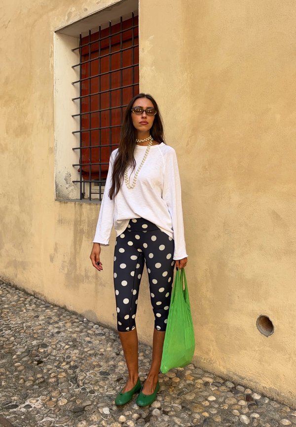 Woman wearing white long-sleeve top and black polka dot leggings stands on cobblestone street holding green bag, next to beige wall with barred window.