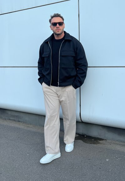 Chaqueta de cremallera negra sobre un suéter azul marino, pantalones beige y zapatillas blancas; de pie contra una pared azul claro con una textura suave.