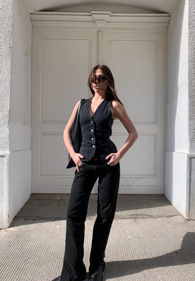 Chaleco de mezclilla negro con un escote en V profundo y botones frontales, combinado con pantalones de campana negros. La modelo sostiene un bolso negro contra una pared neutra.