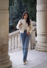 Giacca bianca a trama con bottoni dorati, dolcevita aderente, jeans blue dritti e décolleté beige a punta in un arco di pietra.