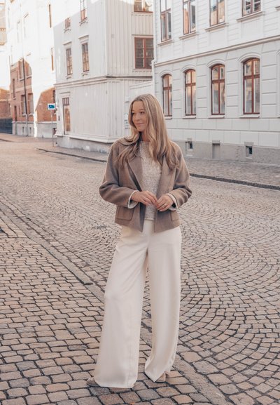 Mujer con largo cabello rubio está de pie en una calle adoquinada vistiendo una chaqueta beige, un suéter claro y pantalones blancos de pierna ancha en un entorno urbano.