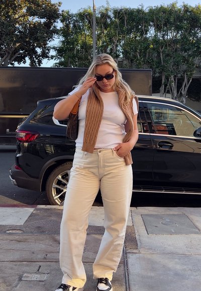 Pantalones beige de pierna ancha, camiseta blanca y un chaleco de punto marrón. Gafas de sol negras y zapatillas en blanco y negro. Fondo urbano con un vehículo oscuro.