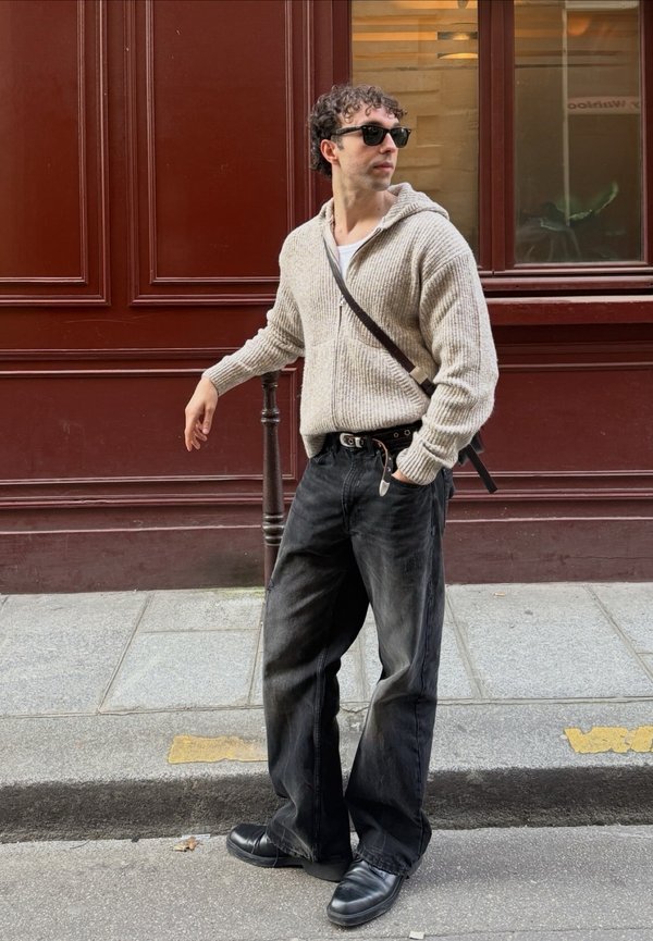 Muškarac sa sunčanim naočalama, bež džemper na patent zatvarač, crne traperice i crne cipele stoji na ulici pokraj crvene fasade zgrade, gledajući bočno.