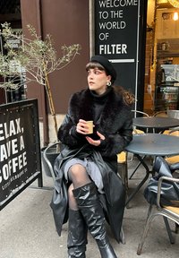 Femme en manteau noir bordé de fourrure et béret assise à l'extérieur d'un café, tenant une petite tasse, regardant sur le côté près des enseignes et des tables du coffee shop.