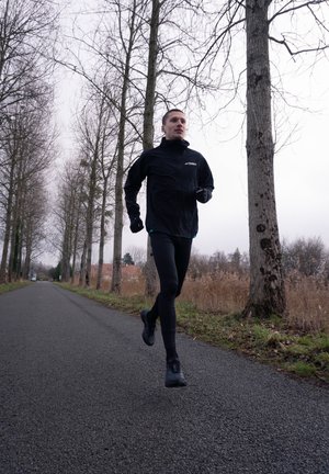 Hombre corriendo en una carretera vacía bordeada de árboles altos y desnudos bajo un cielo nublado, vestido con ropa deportiva negra y guantes.