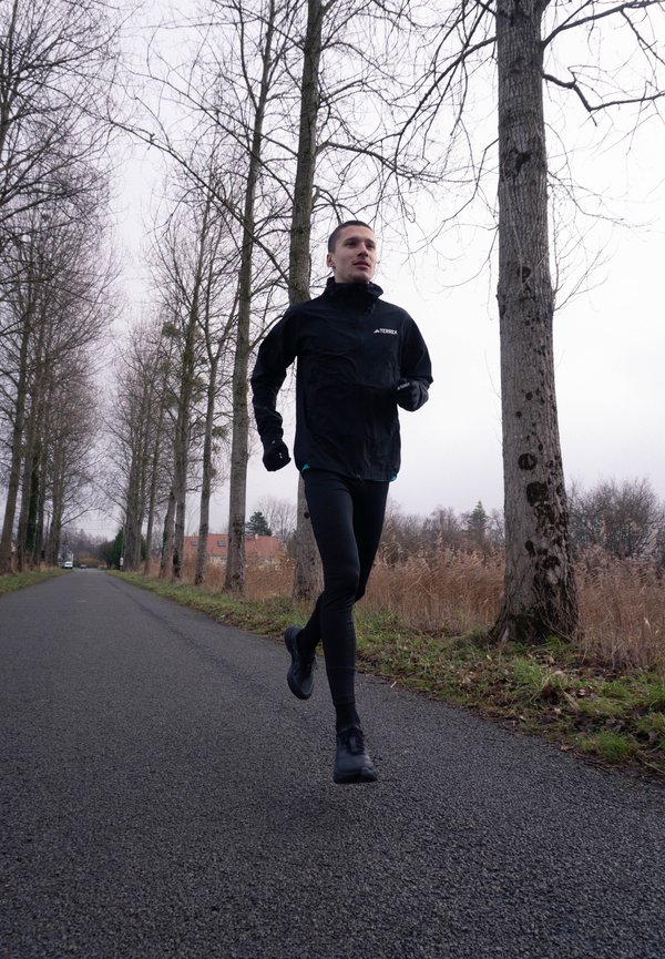 Mann joggt auf leerer Straße, gesäumt von hohen kahlen Bäumen unter bewölktem Himmel, gekleidet in schwarzer Sportbekleidung und Handschuhen.