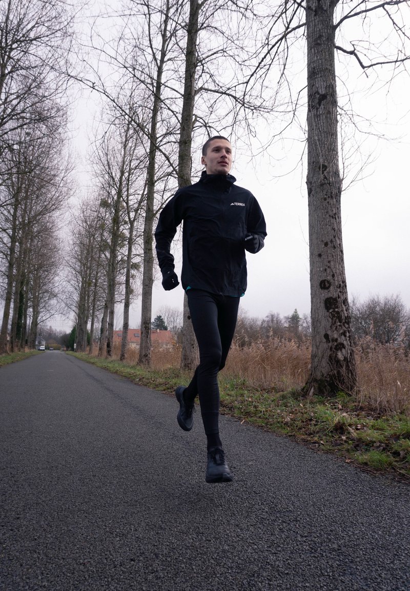 Homme en train de courir sur une route déserte bordée de grands arbres nus sous un ciel couvert, vêtu de vêtements de sport noirs et de gants.