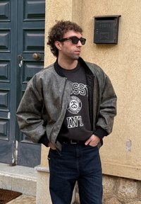 Άντρας με γυαλιά ηλίου και γκρι μπουφάν στέκεται με τα χέρια στις τσέπες κοντά σε ταχυδρομικό γραμματοκιβώτιο στον τοίχο με την επιγραφή "POSTA".