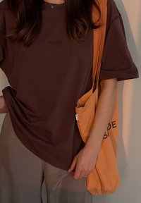 Ruskea ylisuuri t-paita, jossa on kohokuvioitu logo, yhdistettynä oranssiin kangaskassiin, jossa on musta teksti. Silkkinen kangas ja rento istuvuus.