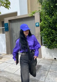 Purple long-sleeve shirt with cutout sleeves, black tie, high-waisted black jeans, and blue cap with text. Model holding a folder.