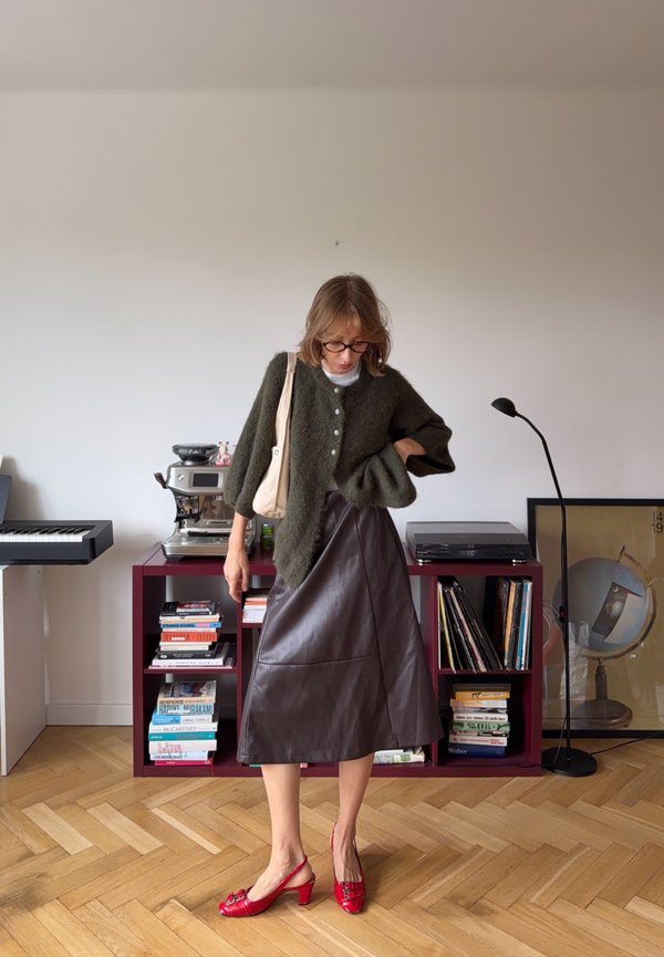 Femme portant un pull vert foncé, une jupe en cuir noire, des talons rouges, des lunettes et un sac beige, debout dans une pièce avec des étagères et un clavier.