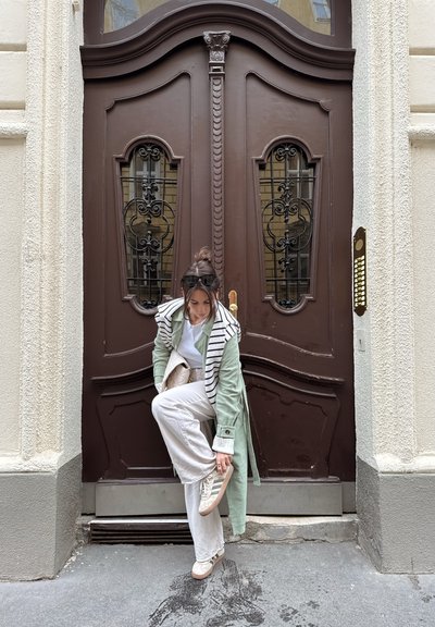 Trench coat texturizado de color verde claro con cuello a rayas, camisa blanca y pantalones crema. Puerta marrón con elaborados hierros negros y herrajes dorados.