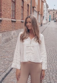 Fehér lenvászon cropped blúz gombos záródással, hosszú ujjakkal és könnyű textúrával, bézs, testhez simuló nadrággal párosítva egy macskaköves utcán.