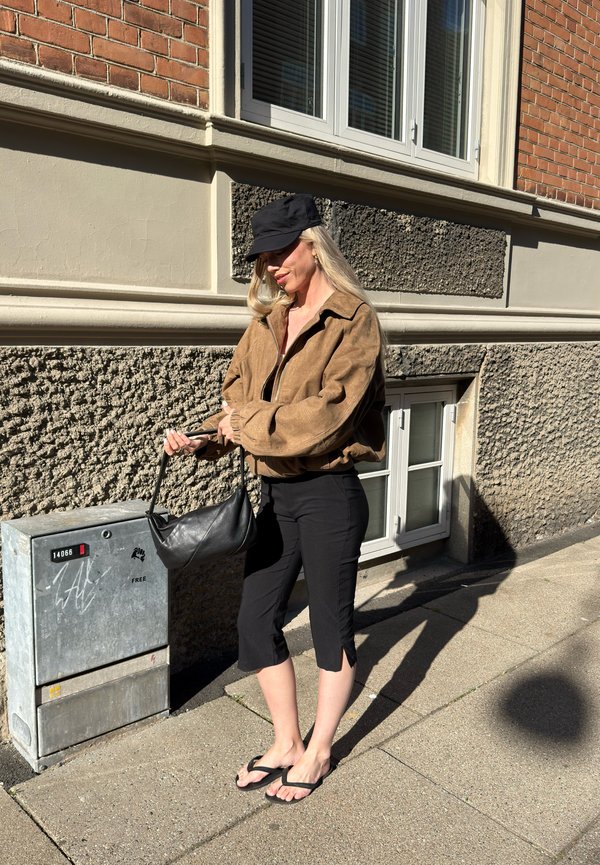 Kvinde iført en brun jakke, sort kasket, sorte korte bukser og flip-flops, der holder en sort håndtaske, mens hun står på en byfortov.