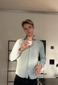 Νεαρός άνδρας με ριγέ πουκάμισο κρατάει ένα λευκό φλιτζάνι, στέκεται σε ένα μίνιμαλ δωμάτιο με φυτά και μια  μαύρη σκάλα-ραφιέρα.