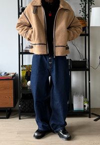 Veste en polaire beige avec des accents marron et une fermeture éclair, associée à un jean en denim bleu foncé à coupe ample. Accessoires comprenant un pendentif rouge.