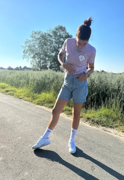 Camiseta lila clara con una etiqueta blanca, pantalones cortos de denim de cintura alta, zapatillas blancas y calcetas blancas hasta la rodilla en una carretera soleada.
