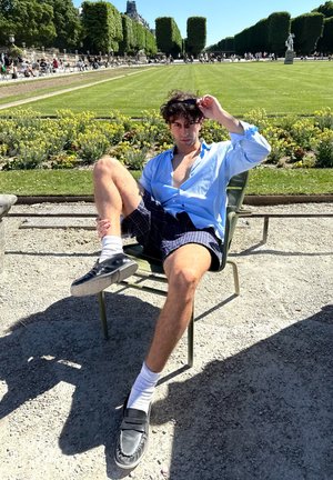 Camisa de manga larga azul claro, pantalones cortos a cuadros negros, calcetines blancos, mocasines negros, sentado en una silla de metal verde en un parque con parterres de flores.