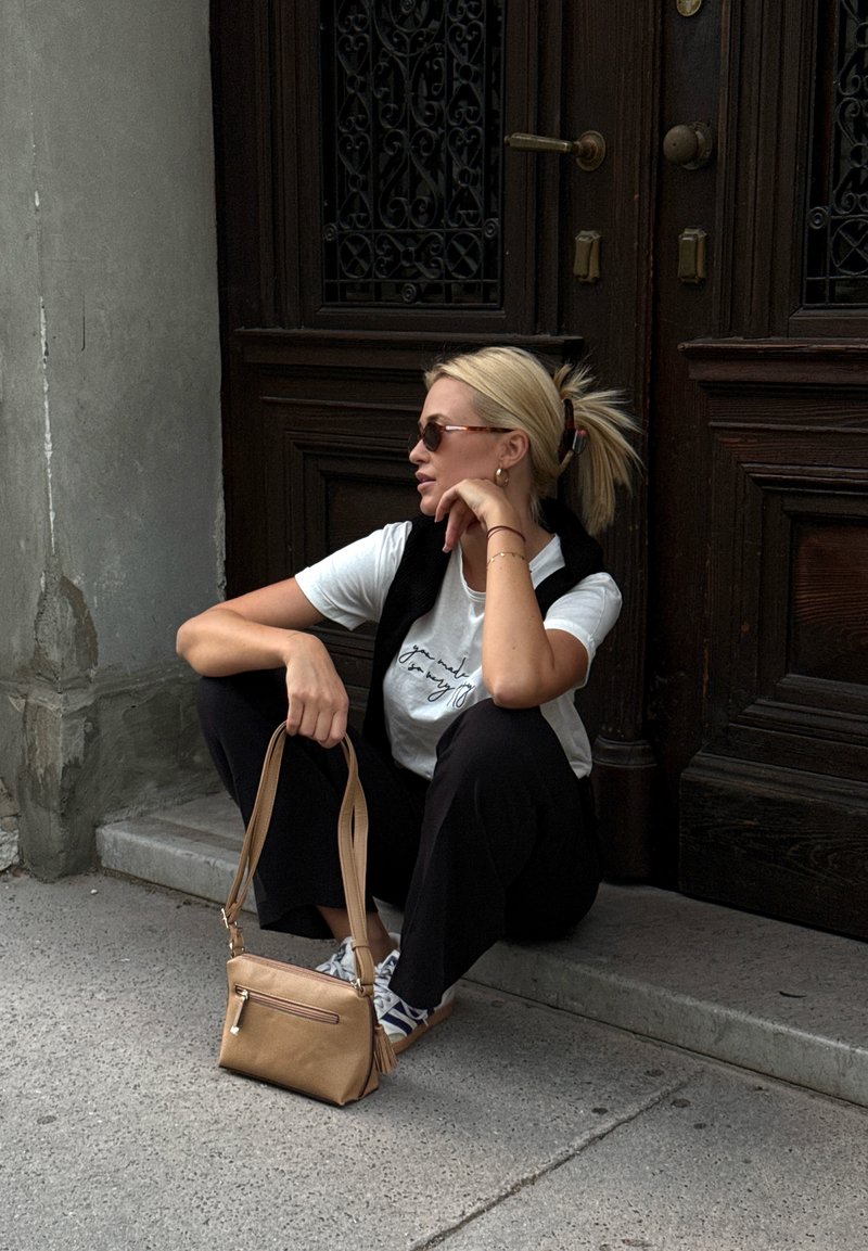 Femme avec des lunettes de soleil assise sur une marche devant une porte en bois sombre, tenant un sac à main beige, portant un pantalon noir et une chemise blanche.
