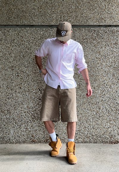 Hombre vestido con una camisa de rayas en rosa claro y pantalones cortos beige, combinada con botas amarillas y calcetines blancos, frente a una pared texturizada.