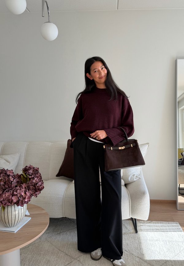 Una mujer con un jersey burdeos y pantalones negros posa en una sala de estar, sosteniendo un bolso marrón oscuro junto a un sofá crema.