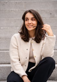 Beżowy sweter z dzianiny z złotymi guzikami, noszony na jasnej bluzce, w połączeniu z czarnymi spodniami. Okulary trzymane w jednej r�ęce. W tle kamienne schody.