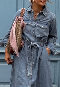 Camicia in denim a vestito con bottoni frontali, due tasche sul petto, cintura in vita e maniche lunghe. Abbinata a una borsa a fantasia e a una sciarpa rosa.