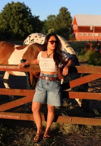 A person leans against a wooden fence, dressed in a white crop top, denim shorts, a brown belt with metal accents, and sandals, with horses in the background.