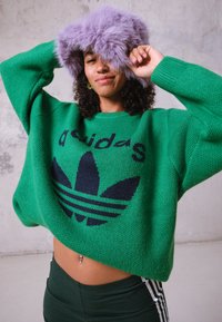 Pulôver oversized verde com um logótipo Adidas azul escuro, combinado com leggings verdes escuras com riscas brancas. Modelo segura um chapéu de lavanda fofinho.