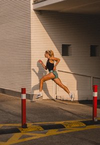 Sportieve shorts in teal, zwart tanktop en witte schoenen. Hardloper in beweging tegen een lichtgekleurde muur en rode veiligheidsbarrières.