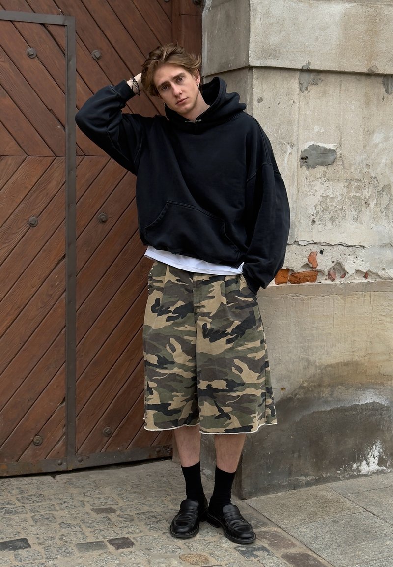 Jeune homme en sweat à capuche noir, short camouflage, chaussettes noires et chaussures, debout dans une rue pavée devant une porte en bois et un mur de pierre ancien.