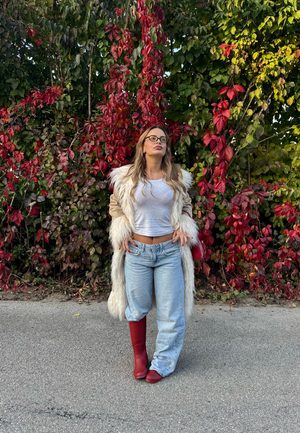 Umelá kožušina s béžovým telom a bielym lemovaním, šedý crop top, svetlomodré široké džínsy a červené kozačky po kolená na pozadí červenej a zelenej zelene.