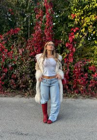 Umelá kožušina s béžovým telom a bielym lemovaním, šedý crop top, svetlomodré široké džínsy a červené kozačky po kolená na pozadí červenej a zelenej zelene.