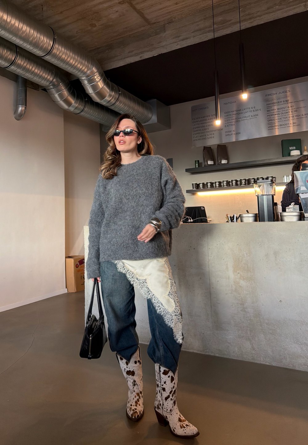 Femme portant un pull gris, un jean orné de dentelle et des bottes à motif vache tenant un sac noir à l'intérieur d'un café moderne au décor industriel.