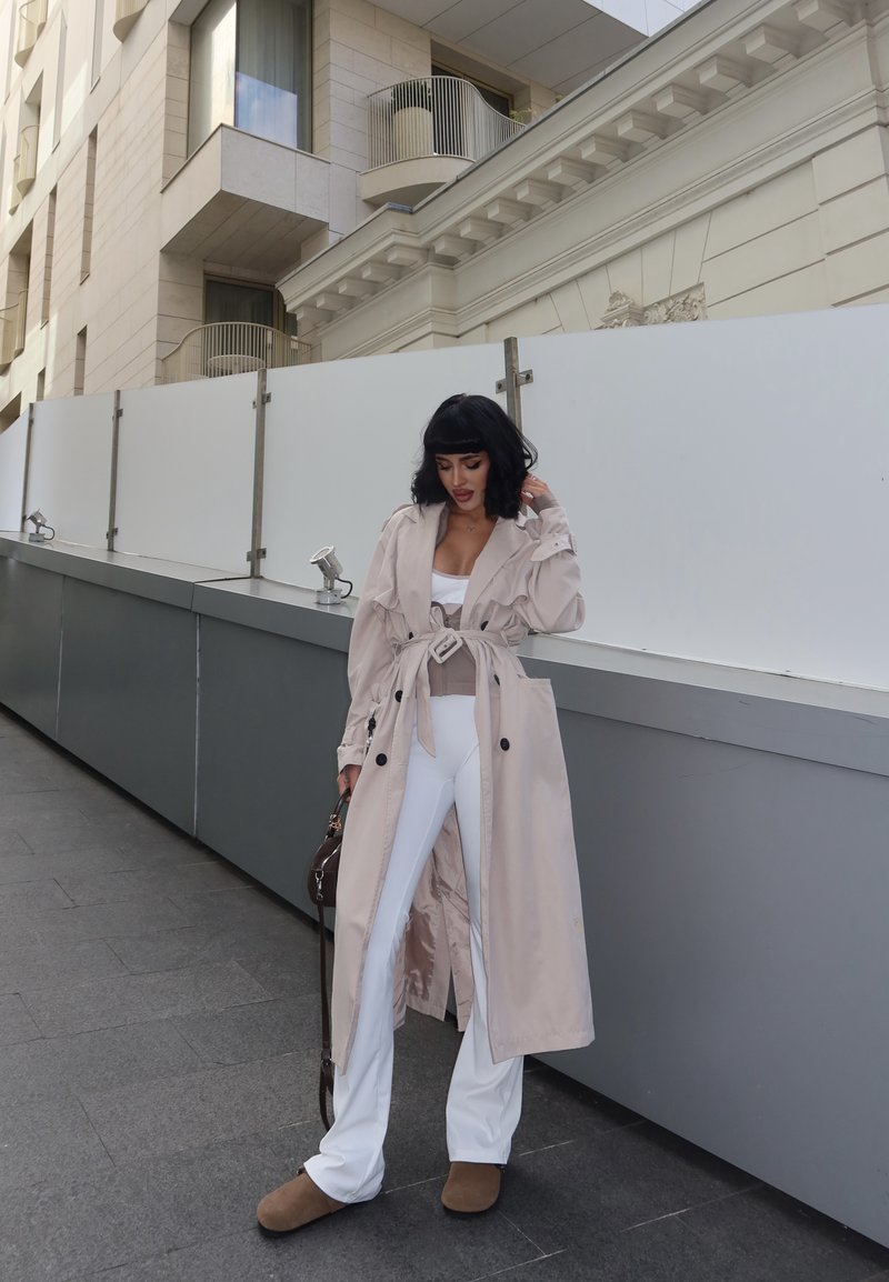 Manteau beige avec ceinture, manches longues et boutons sur un haut blanc ajusté et un pantalon blanc évasé. Chaussures brunes et petit sac à main.