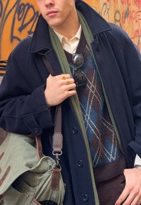 Marineblauer Wollmantel, kombiniert mit einem grünen Schal, einem braunen Argyle-Pullover und einem weißen Hemd. Accessoires sind Sonnenbrillen und Ringe.