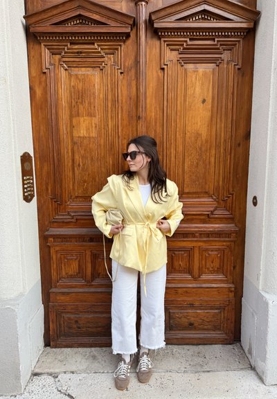 Mujer con blazer amarillo y pantalones blancos, sosteniendo un bolso beige, parada frente a una puerta doble de madera ornamentada.