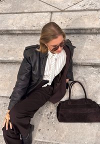 Svart läderjacka, randig vit skjorta, bruna byxor, mörka solglasögon och en mörk mocka handväska på stentrappor.