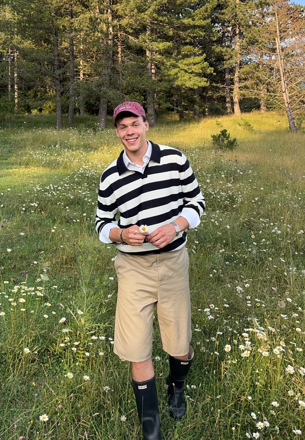 Jeune homme en chemise rayée, short beige, bottes noires et casquette rouge souriant en tenant une marguerite dans un champ herbeux avec des fleurs sauvages et des arbres.