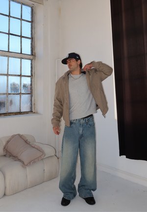 Hombre con chaqueta beige, camiseta gris, vaqueros holgados y gorra negra, de pie en el interior junto a una ventana grande y un sofá blanco con cojines.