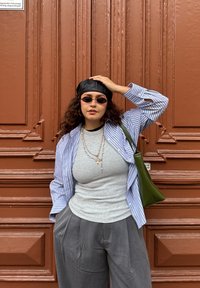 Chemise oversize à rayures bleues et blanches, haut gris ajusté, pantalon gris ample, casquette en cuir noire, sac à main vert, et lunettes de soleil foncées devant un mur marron.